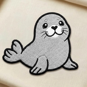 May include: Embroidered patch featuring a gray seal with a black outline. The seal has a smiling face with a heart-shaped nose and whiskers. The patch is designed to be ironed or sewn onto fabric, suitable for clothing or accessories.