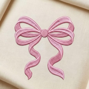 Könnte beinhalten: Nahaufnahme einer rosa gestickten Schleife auf cremefarbenem Stoff. Die Schleife hat detaillierte Nähte und fließende Bänder, die ein zartes Design schaffen. Die Schleife ist zentriert und zeigt ihre symmetrische Form und komplizierten Details.