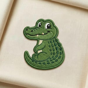 May include: Embroidered green alligator patch with a smiling face and white accents. The patch features detailed stitching and a friendly design, perfect for adding to clothing or accessories.