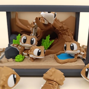 Może przedstawiać: Diorama przedstawiająca kreskówkowe jaszczurki w scenerii pustynnej. Jaszczurki są brązowo-białe z dużymi oczami. Scena obejmuje element wodny, skały i duży pień drzewa. Diorama jest zamknięta w szarej ramie.