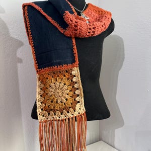May include: A handmade scarf with a navy blue body and orange crochet trim. The scarf features a square crochet design in shades of tan and brown, with orange fringe. The scarf is displayed on a black mannequin.