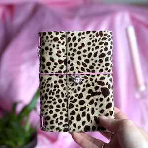 May include: A rectangular notebook covered in a tan and dark brown leopard print fur texture. The notebook is secured by a bright pink elastic band featuring a silver-toned metal charm shaped like a heart with wings and a central clear crystal.