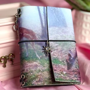 Puede incluir: Un diario rectangular con una impresión fotográfica de un ciervo en un bosque brumoso. La cubierta está asegurada con una banda elástica negra adornada con un dije plateado en forma de estrella. Se aprecian ojales metálicos plateados.