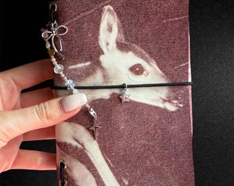 Może przedstawiać: Prostokątna okładka na notatnik z fotograficznym nadrukiem jelenia w odcieniu sepii. Czarna gumka owija środek, ozdobiona sznurkiem z koralikami, srebrzystą kokardką, zawieszką w kształcie gwiazdki oraz zawieszką w kształcie czteroramiennego krzyża.