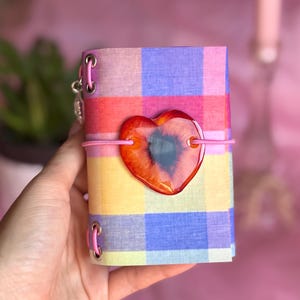 May include: A small notebook featuring a colorful plaid pattern in shades of purple, pink, yellow, and orange. The cover is secured with a pink elastic band holding a large, glossy, heart-shaped resin charm with red and black swirling colors.