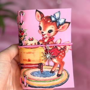 May include: A pink notebook featuring a vintage illustration of a fawn wearing a red polka dot dress and a blue bow. The cover is bound with pink elastic cord through silver metal eyelets and includes a silver star-shaped charm on the front.