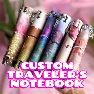 May include: A collection of six traveler's notebooks featuring various printed designs, including floral, landscape, and abstract patterns. Each notebook is bound with metal eyelets, silver-toned charms, and elastic cords in pink, black, or white.