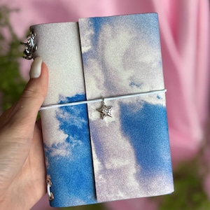 May include: A small notebook with a cover featuring a blue sky and white cloud print. The notebook is secured by a white elastic band that holds a silver-toned star charm. A silver-toned metal ring is attached to the side of the notebook cover.