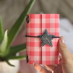 May include: A small rectangular notebook featuring a pink and white gingham pattern. The cover is secured by a black elastic band that wraps around the center, holding a five-pointed star ornament with a black and white spiral design.