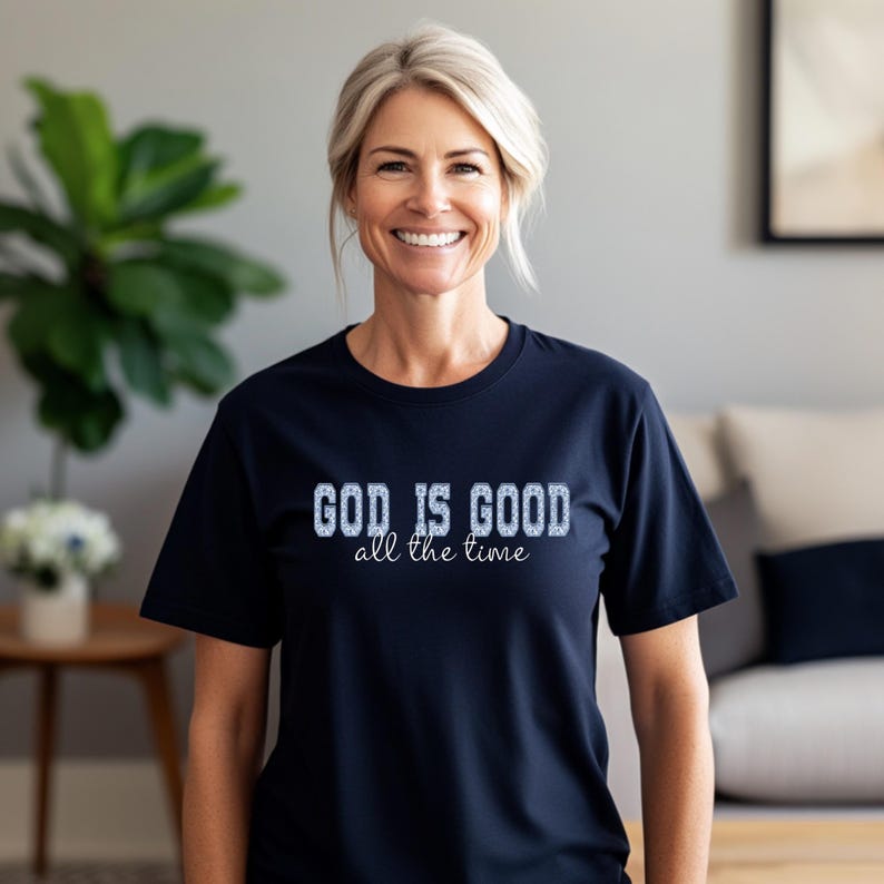 Navy blue t-shirt with the text "GOD IS GOOD all the time" in a textured, light blue font. The shirt is short-sleeved and has a crew neck. The image shows a person wearing the shirt.