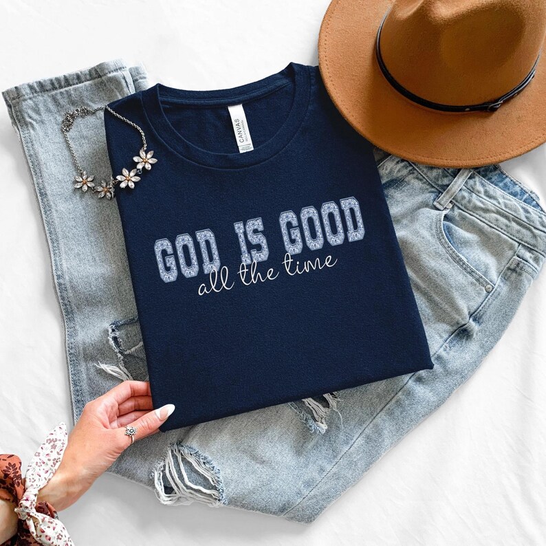 Navy blue crew neck t-shirt with the text "GOD IS GOOD all the time" in a white, textured font. The shirt is paired with light wash denim jeans and a brown hat.