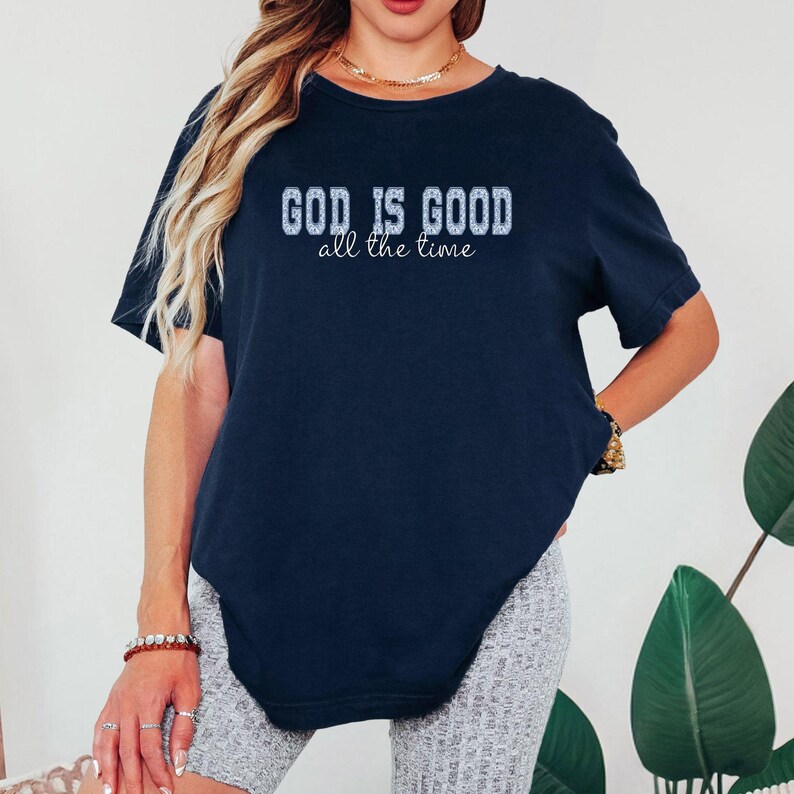 May include: Navy blue t-shirt with the text "GOD IS GOOD all the time" in a white, textured font. The shirt has short sleeves and a relaxed fit. The overall style is casual and comfortable.
