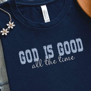 Navy blue t-shirt with the text "GOD IS GOOD all the time" in a decorative font. The words are in a light blue, textured design. A silver necklace with flower-shaped pendants is visible in the upper left corner.