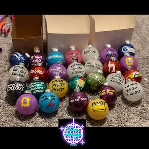 Puede incluir: Una colección de adornos navideños brillantes con varias citas y frases de la serie de televisión Friends. Los adornos son redondos y coloridos, con texto en blanco y negro. Algunos tienen imágenes de los personajes y objetos icónicos del programa.