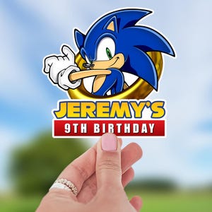 May include: A colorful sticker featuring Sonic the Hedgehog, with blue spikes, a white glove, and a golden ring. The sticker includes the text "JEREMY'S 9TH BIRTHDAY" in yellow and red. The sticker is held by a person.
