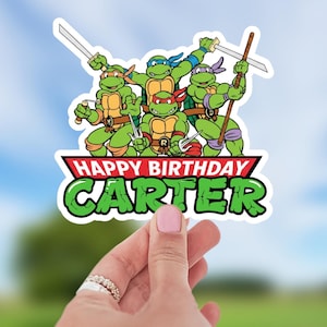 May include: A sticker featuring the Teenage Mutant Ninja Turtles with weapons. The sticker has the text "HAPPY BIRTHDAY CARTER" in green and red. The turtles are green with different colored masks.