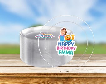 May include: A stack of clear plastic plates with a transparent plate on top. The plates have a sticker that says "Happy Birthday Emma" with cartoon characters. The plates are on a wooden surface.
