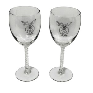 May include: Two clear glass wine glasses with twisted stems. Each glass features a black printed graphic of a scimitar, a pharaoh head, and a star, with the text ABU BEKK, 1999, and ALAN JENSEN POTENTATE 1999 printed on the bowl of the glass.