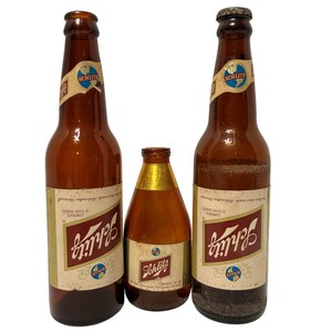 May include: Three amber glass Schlitz beer bottles of varying sizes. Two tall bottles feature white labels with red Schlitz branding, globe logos, and 12 fluid ounces text. A smaller, shorter bottle has a gold neck and a similar Schlitz label.
