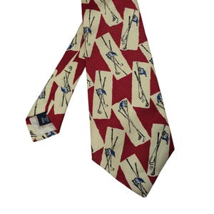 May include: A necktie featuring a repeating pattern of crossed golf clubs and blue golf flags with the number 8 on a cream-colored background, set against diagonal red stripes. The tie includes a small black label reading Made By Hand.