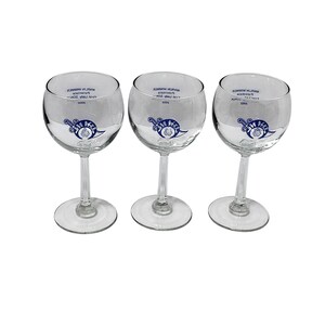 May include: Three clear glass wine glasses with stems and round bases. Each glass features a blue circular logo with a sword and text, plus the printed text MARLIN WIMMER, Potentate, First Lady SONA, 2002. The glasses are identical in design and shape.