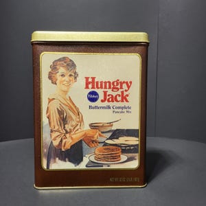 May include: A rectangular brown metal tin of Hungry Jack Buttermilk Complete Pancake Mix. The label features an illustration of a woman cooking pancakes. The text "Hungry Jack" is in red, with "Pillsbury" in blue. The net weight is 32 oz (2 lb) 907g.