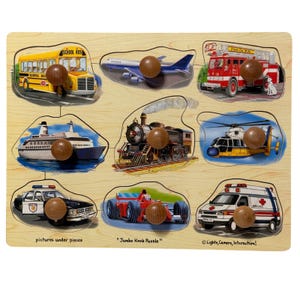 May include: A wooden peg puzzle featuring nine transportation vehicles including a yellow school bus, airplane, red fire truck, cruise ship, steam train, helicopter, police car, race car, and ambulance, each with a round wooden knob for grasping.