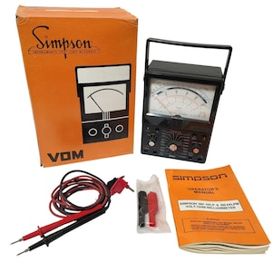 May include: An orange box with the Simpson logo and the text "INSTRUMENTS THAT STAY ACCURATE" next to a black VOM meter. A black meter with a handle, red and black wires, and an orange manual are also present.