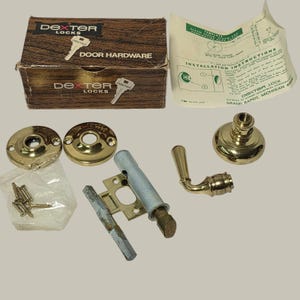 May include: A Dexter Locks door hardware set including a brown cardboard box, installation instructions, two circular brass rosettes, a brass lever handle, a metal latch mechanism, a square spindle, and a small plastic bag containing mounting screws.