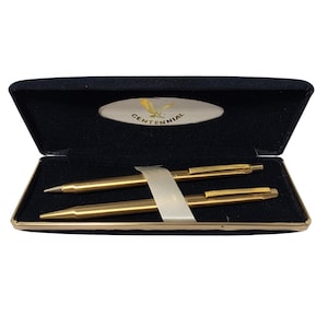 May include: A set of two gold-toned pens secured by a white band inside a black velvet-lined rectangular case. The interior lid features an oval white emblem with a gold eagle graphic and the text CENTENNIAL printed in black capital letters.