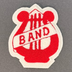 May include: A red and white embroidered patch with the word "BAND" in white letters. The patch features a musical instrument design with a white border. The patch is likely for a band or musical group.