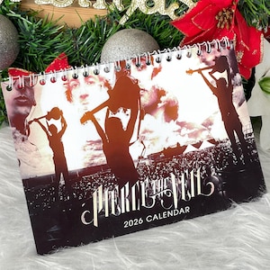 Könnte beinhalten: Ein Spiral-Kalender 2026 mit der Band Pierce the Veil. Das Cover zeigt Bandmitglieder und Silhouetten von Musikern, die Gitarre spielen. Der Kalender ist vor dem Hintergrund einer Konzertmenge platziert.