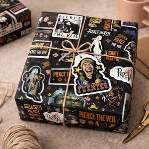 May include: A wrapped gift box with black paper decorated with Pierce the Veil stickers and text. The box is tied with twine and a tag featuring a band member. The theme is music and band merchandise.