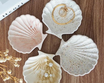 Puede incluir: Cuatro cuencos en forma de concha en tonos blanco, beige y rosa, sobre una superficie de madera. Uno contiene una pulsera y anillos dorados, otro pendientes de perlas. Los cuencos varían en textura y patrón.