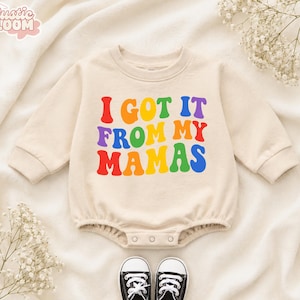May include: A beige long-sleeve baby bodysuit featuring the colorful text I GOT IT FROM MY MAMAS in a wavy font. The garment has a crew neckline, ribbed cuffs, and a snap-button closure at the bottom. A pair of black and white sneakers is included.