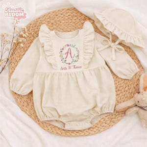 May include: Cream-colored baby romper and matching bonnet set. The romper features a floral embroidered initial "A" and the name "Anita W. Thomas". The set includes a long-sleeved romper with ruffle trim and a bonnet with a bow.