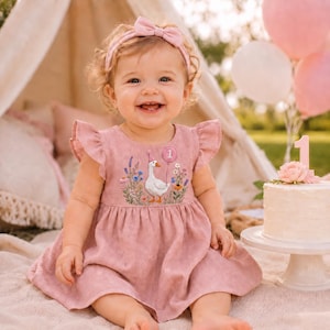 May include: A dusty rose-colored dress with flutter sleeves and a headband featuring a matching bow. The dress bodice displays an embroidered scene of a white goose wearing a party hat, surrounded by colorful wildflowers and a balloon with the number 1.
