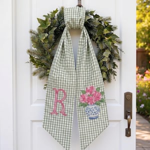 May include: A green and white gingham door scarf with a pink embroidered letter "R" and a floral design. The scarf is hanging on a white door with a wreath.