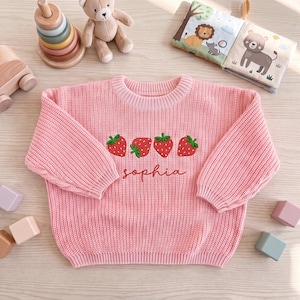 May include: A light pink long-sleeve knit sweater featuring a row of four embroidered red strawberries with green leaves across the chest. Below the strawberries, the name "sophia" is embroidered in a cursive red script font on the front.