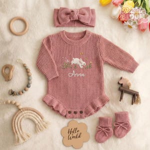May include: A pink knitted baby outfit set, including a romper with a bunny embroidery, a headband, and socks. The romper has the name "Anna" embroidered on it. A wooden sign says "Hello World".