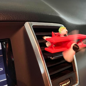 May include: A red airplane-shaped car air vent clip featuring a cartoon character in a pilot's helmet. The propeller is blurred, indicating motion. The accessory is placed on a car's dashboard near the air vent.