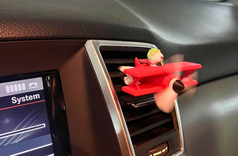 May include: A red airplane-shaped car air vent clip featuring a cartoon character in a pilot's helmet. The propeller is blurred, indicating motion. The accessory is placed on a car's dashboard near the air vent.