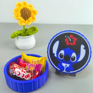 K&ouml;nnte beinhalten: Ein blauer, runder Beh&auml;lter gef&uuml;llt mit verpackten S&uuml;&szlig;igkeiten, neben einem blauen und schwarzen Stitch-Charakterdesign. Eine kleine, gelb geh&auml;kelte Sonnenblume in einem wei&szlig;en Topf ist ebenfalls vorhanden.