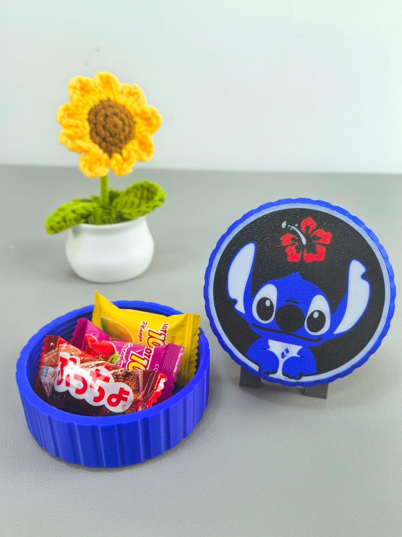 K&ouml;nnte beinhalten: Ein blauer, runder Beh&auml;lter gef&uuml;llt mit verpackten S&uuml;&szlig;igkeiten, neben einem blauen und schwarzen Stitch-Charakterdesign. Eine kleine, gelb geh&auml;kelte Sonnenblume in einem wei&szlig;en Topf ist ebenfalls vorhanden.