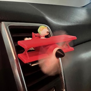 May include: A red biplane car air freshener featuring a cartoon character pilot with a green helmet and goggles. The air freshener is placed in a car vent, with a white vapor cloud coming from the plane.