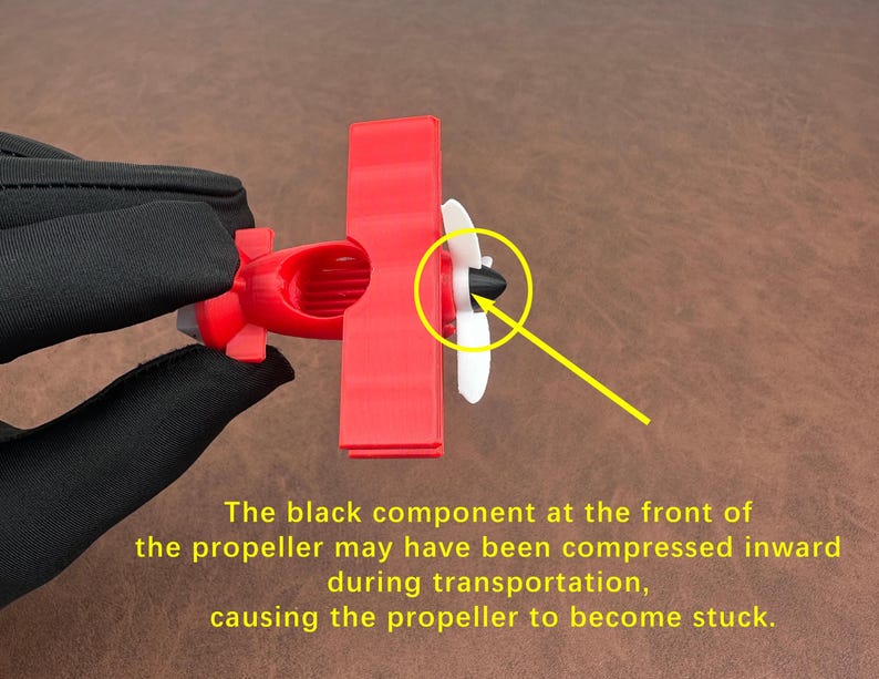 May include: A small red toy airplane with a white propeller and a black component at the front. The text on the image says the propeller may be stuck due to compression during transportation.