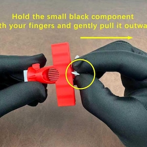 May include: A close-up of a person's hands wearing black gloves, holding a red plastic component. The text in the image reads: "Hold the small black component with your fingers and gently pull it outward."