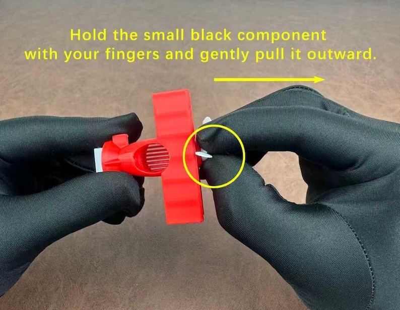 May include: A close-up of a person's hands wearing black gloves, holding a red plastic component. The text in the image reads: "Hold the small black component with your fingers and gently pull it outward."