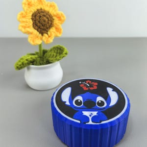 K&ouml;nnte beinhalten: Ein blauer Beh&auml;lter mit einem schwarzen Deckel mit einem Cartoon-Charakter-Design. Im Hintergrund befindet sich eine geh&auml;kelte Sonnenblume in einem wei&szlig;en Topf. Die Sonnenblume hat gelbe Bl&uuml;tenbl&auml;tter und eine braune Mitte. Der Beh&auml;lter hat eine gerippte Textur.