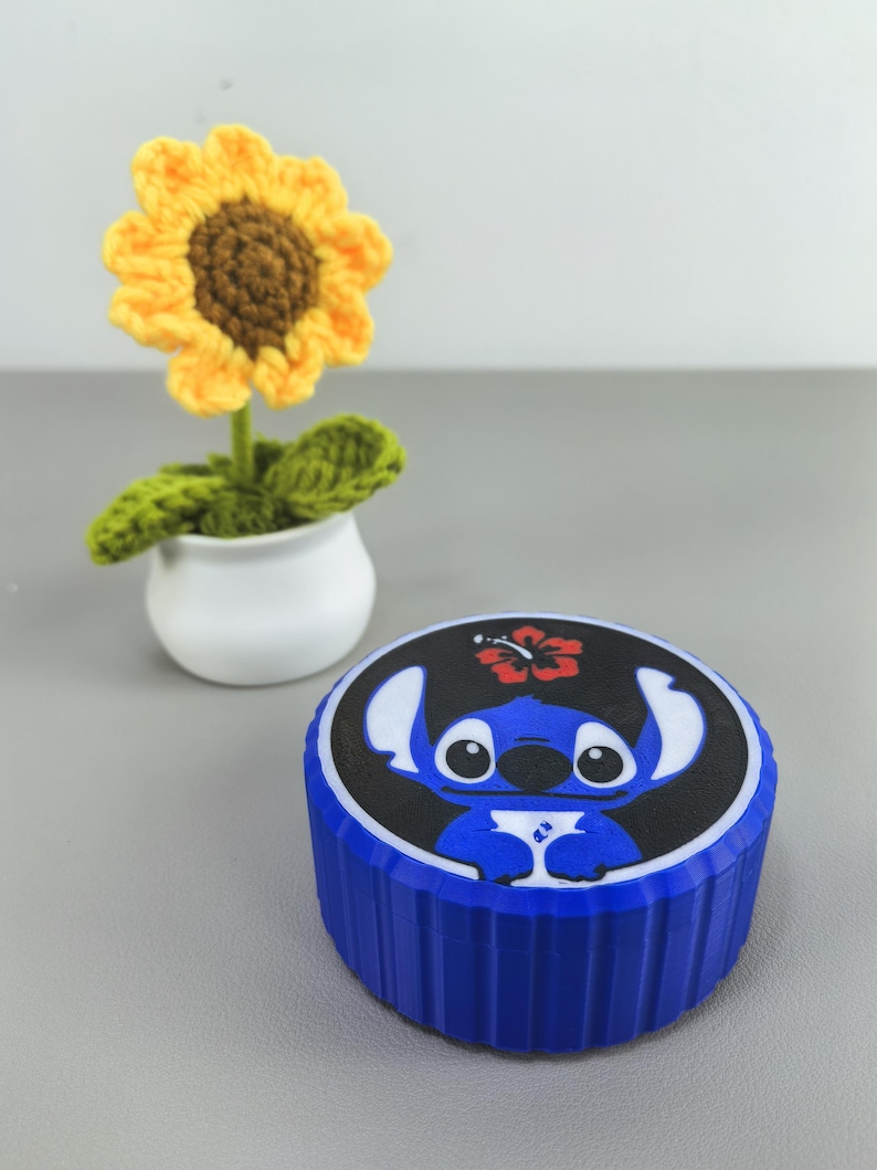 K&ouml;nnte beinhalten: Ein blauer Beh&auml;lter mit einem schwarzen Deckel mit einem Cartoon-Charakter-Design. Im Hintergrund befindet sich eine geh&auml;kelte Sonnenblume in einem wei&szlig;en Topf. Die Sonnenblume hat gelbe Bl&uuml;tenbl&auml;tter und eine braune Mitte. Der Beh&auml;lter hat eine gerippte Textur.